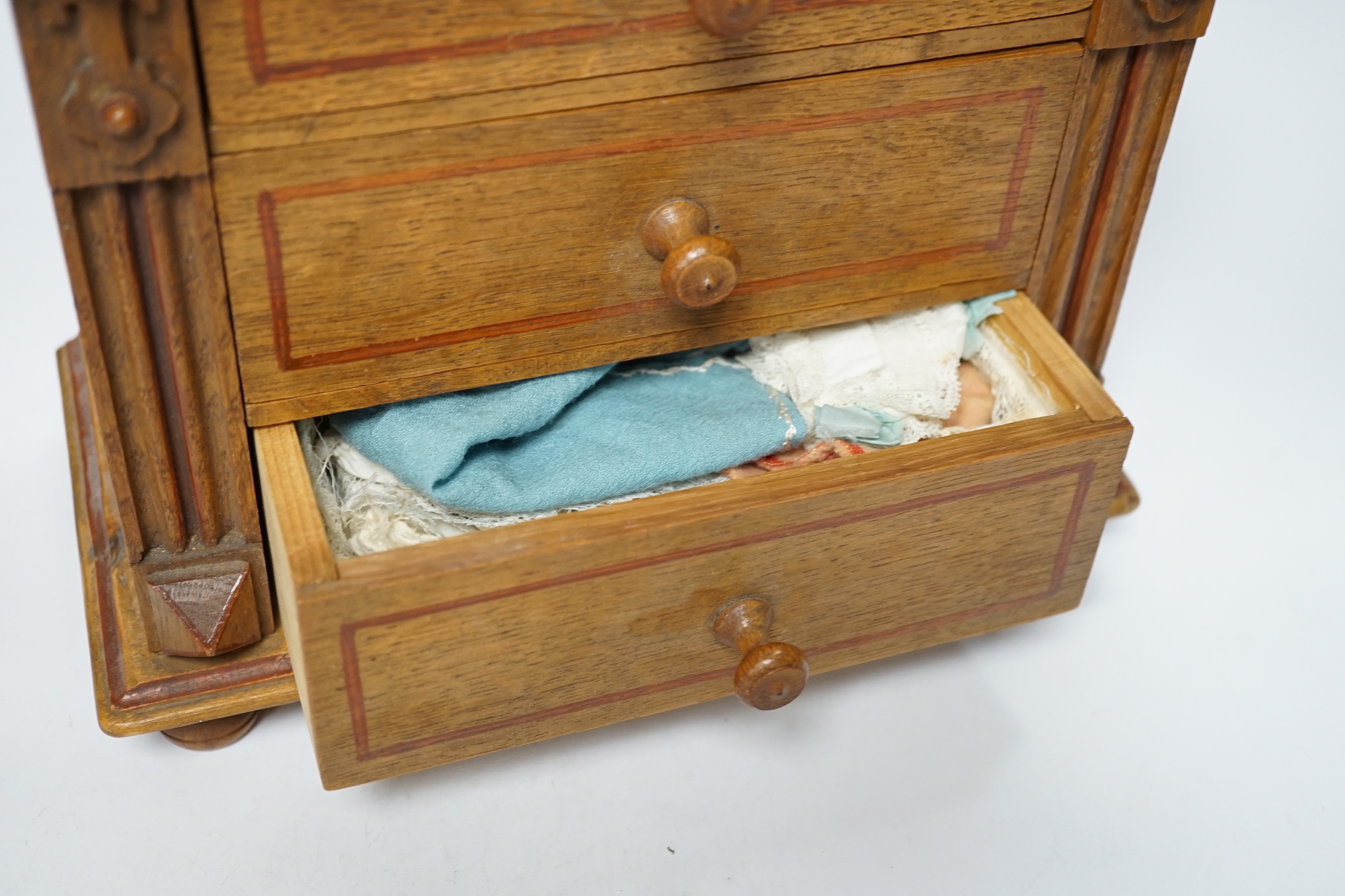 A miniature oak chest of drawers and contents (miniature bisque headed dolls and linen) height 22cm